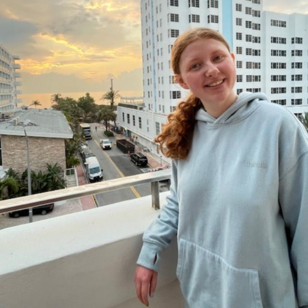 Jordan is posing and smiling on an apartment balcony with a beautiful sunset in the background.