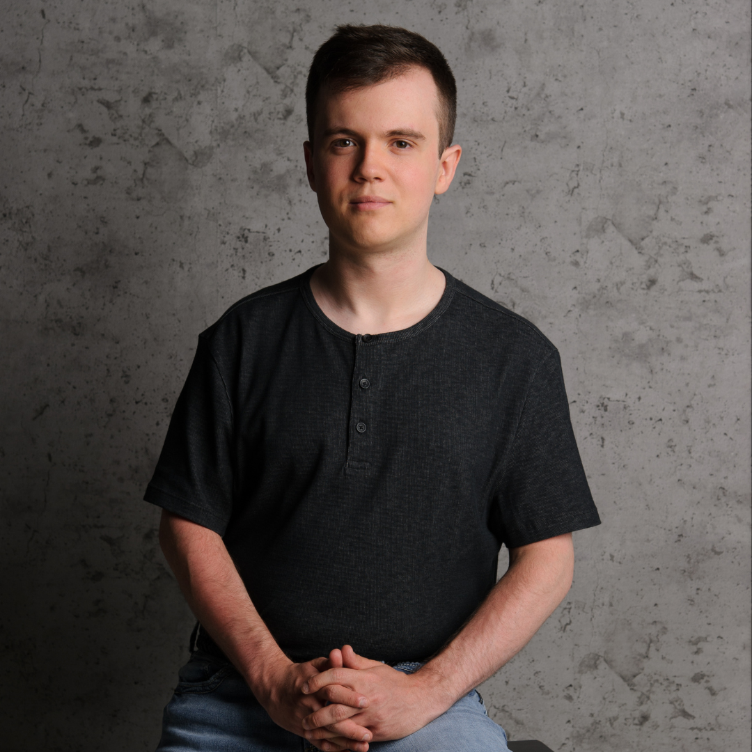 Logan poses in a grey shirt and blue jeans, he is sitting on a tall stool in front of a dark grey background.