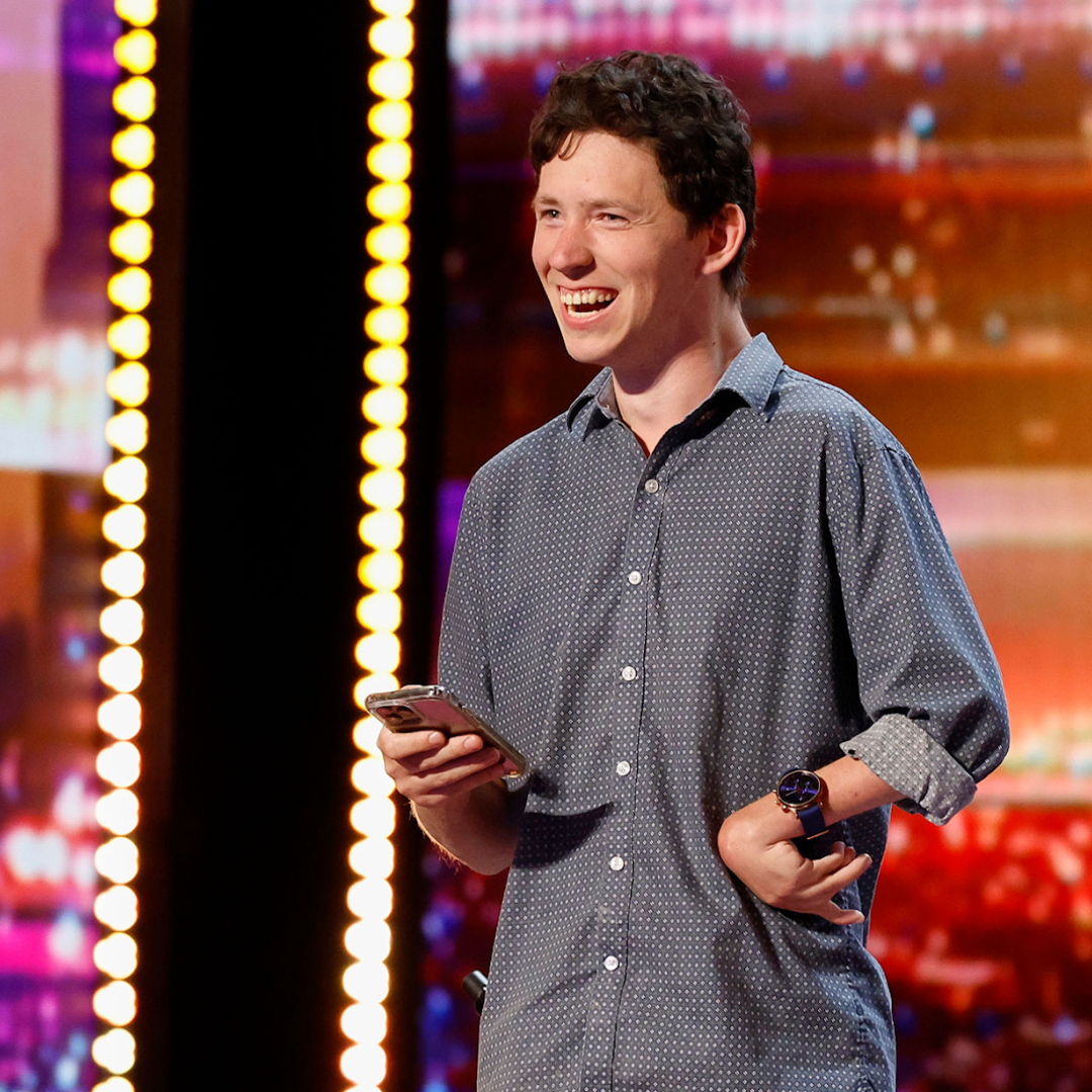 Ahren is on stage performing comedy on the hit tv show, America's Got Talent. He is smiling at the crowd, holding his cellphone, which he uses to communicate.