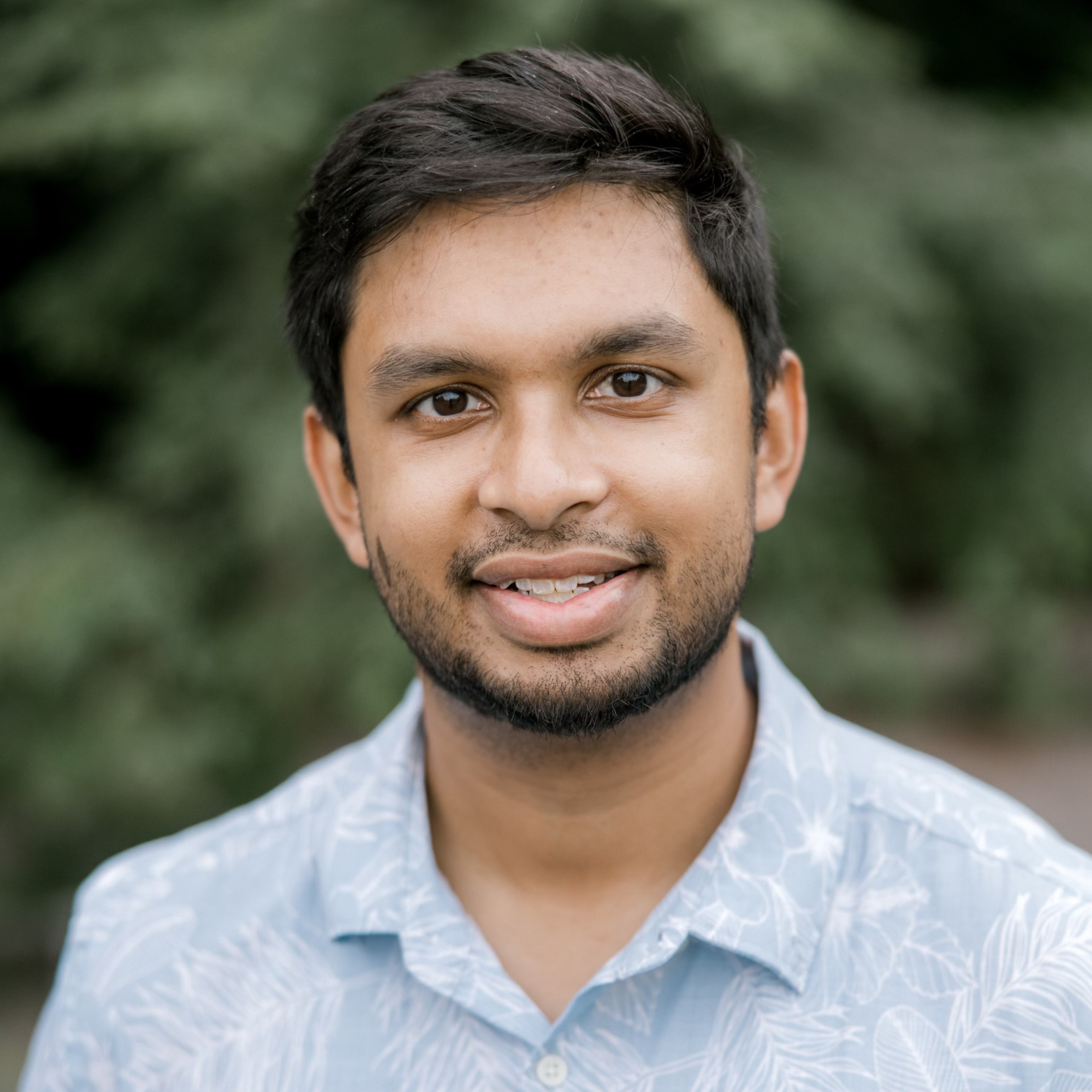 Rageen is standing outside as he smiles for a headshot. The green forest is blurred in the background.