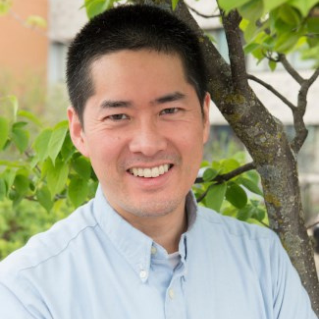 Michael Cheng is standing outside on a sunny day smiling for his headshot, the green outdoor background is blurred behind him.