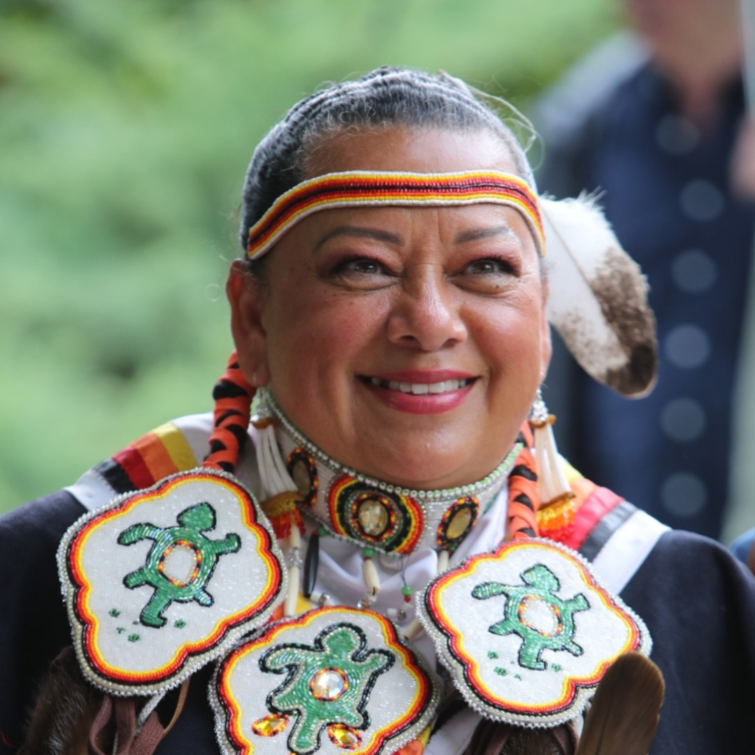 Anishinaabe Traditional Grandmother, Kim Wheatley, is lookingjust off camera and smiling. There is a green forest blurred in the background.