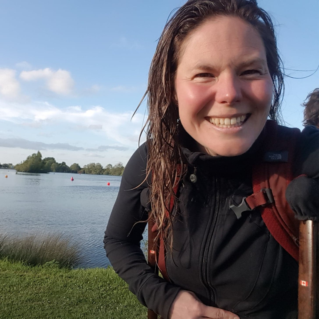 Stephanie is white with long brown hair. They are in a black sweater, smiling for a selfie in front of a beautiful lake and blue sky.