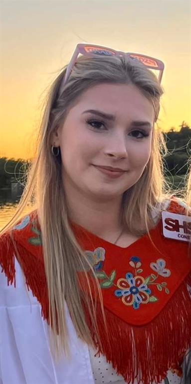 A girl stands smiling at the camera, she is standing in front of a lake and a beautiful sunset.