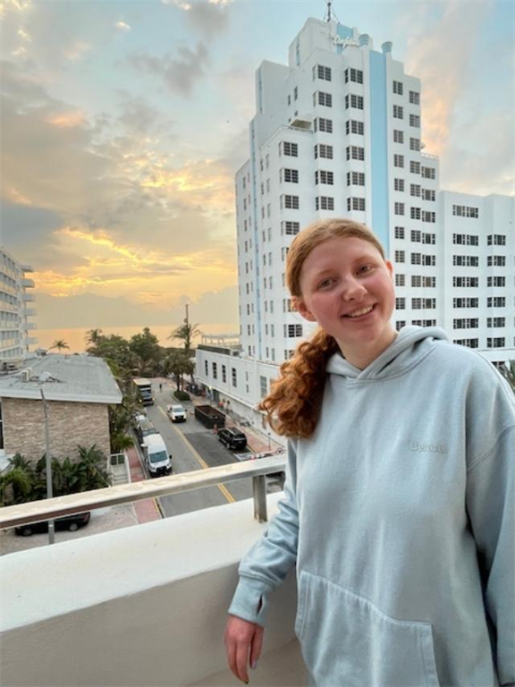 Jordan is posing and smiling on an apartment balcony. The sky behin her is a beautiful sunset.