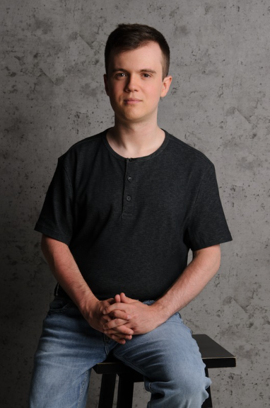 Logan is sitting on a stool, posing for a photo. He is wearing jeans and a black shirt with a dark grey back drop.